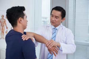 Horizontal portrait of mature Asian traumatologist wearing white coat examining sporty young man palpating his knee