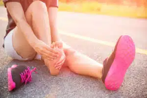 A woman is holding her foot due to pain on the top while sitting on the road with a pair of her running shoes off.