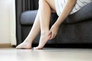 A woman is sitting on a sofa, holding her foot due to pain from flat feet.